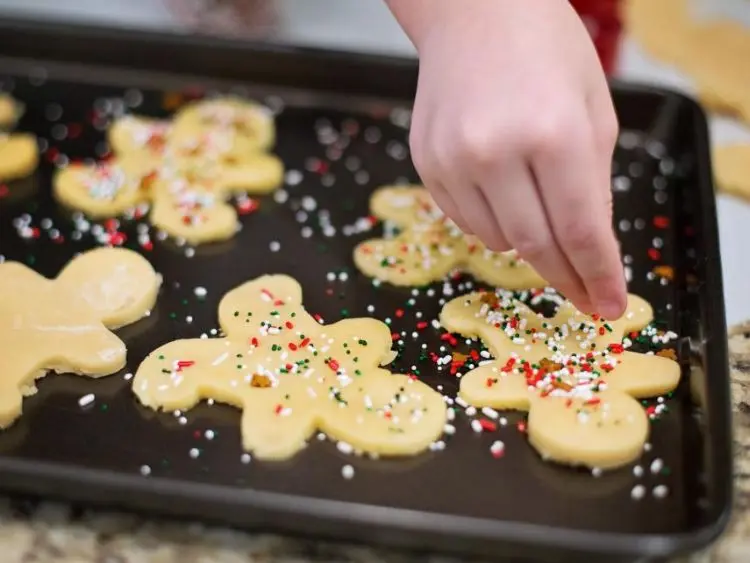 The image shows a close-up of someone's hand sprinkling colorful sprinkles onto uncooked cookies that are shaped like gingerbread men.