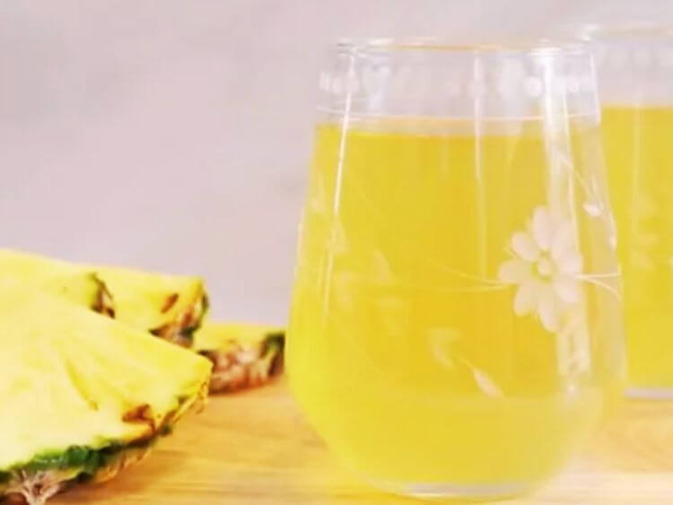 The image shows a glass of pineapple juice next to a pineapple slice.