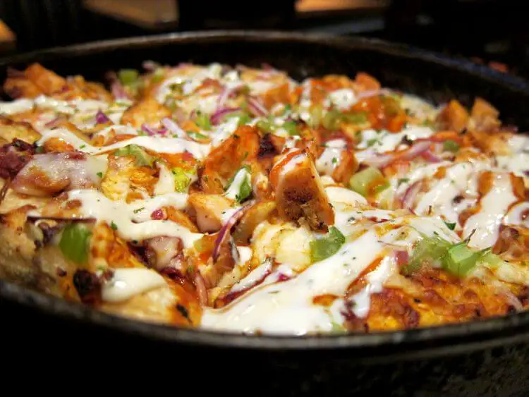 The image shows a buffalo chicken flatbread pizza, freshly baked and presented in a black pan.