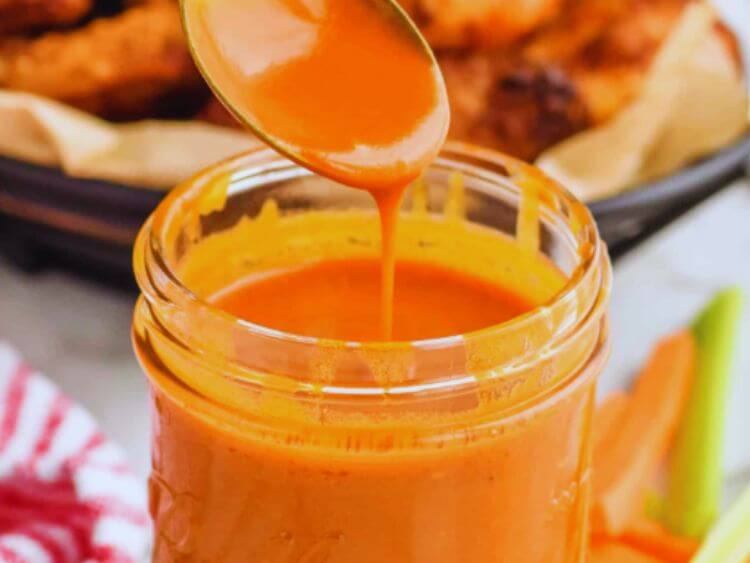 The image shows a smooth, creamy Buffalo sauce with a bright, orange hue. It is being poured from a spoon, demonstrating its thick yet pourable consistency, which coats the spoon evenly—a sign of a well-emulsified sauce.