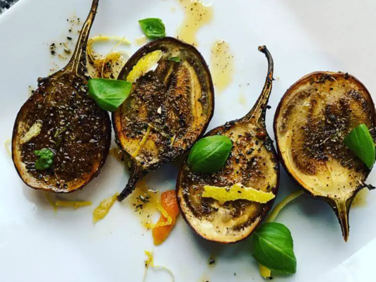 The image displays a plate of grilled baby eggplants. Each eggplant half has its stem intact, suggesting they have been halved lengthwise.