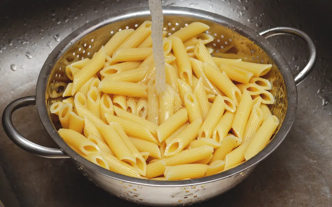 Metal strainer filled with pasta being rinsed under running water from a kitchen faucet.