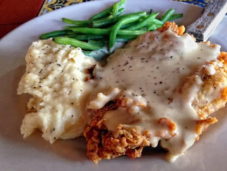 The image depicts a plate of food that includes a serving of chicken-fried steak covered in creamy white gravy peppered with black spots, which is likely a peppered country gravy.