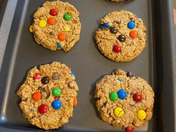 a baking tray with four large, thick cookies, likely gluten-free monster cookies given their hearty appearance and mix of ingredients visible.